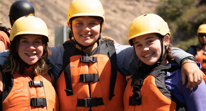 three outward bound students wearing life jackets and helmets wrap their arms around each other's shoulders and pose for the camera 